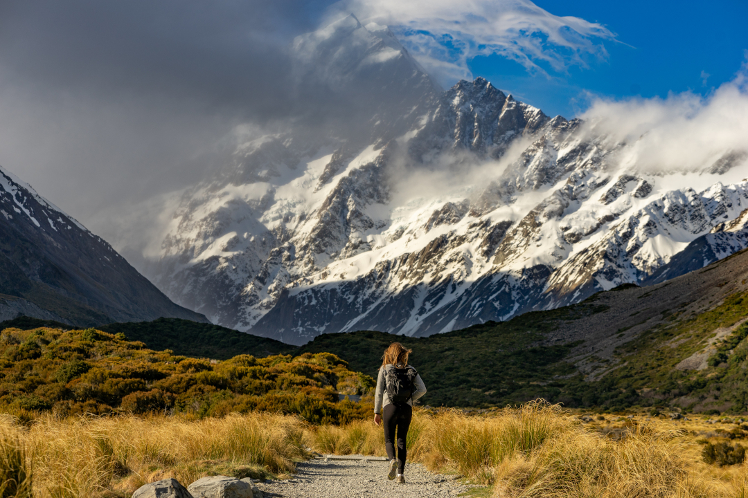 A woman hiking down a trail that leads to a massive mountain in the distant background.